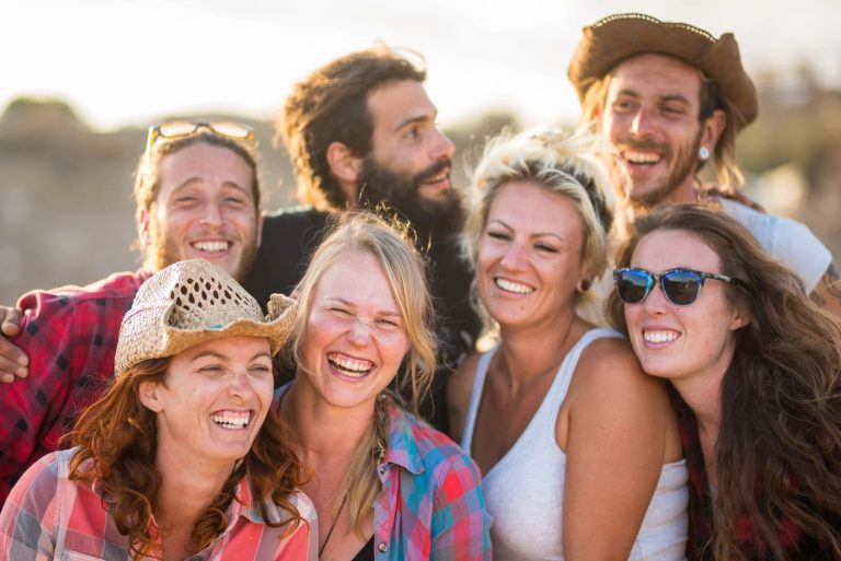 A large, diverse group of smiling friends poses closely together for a candid photo outdoors during the day.