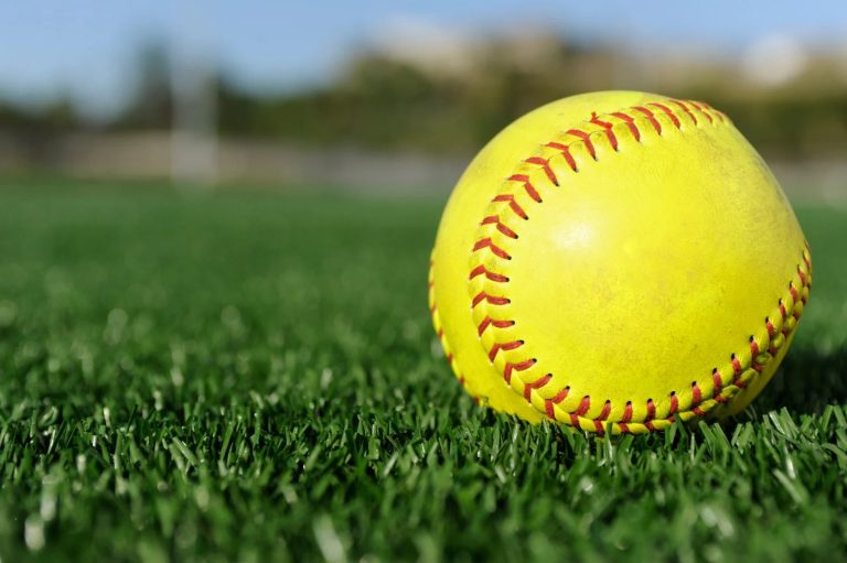 A yellow softball laying on green turf on a sunny day