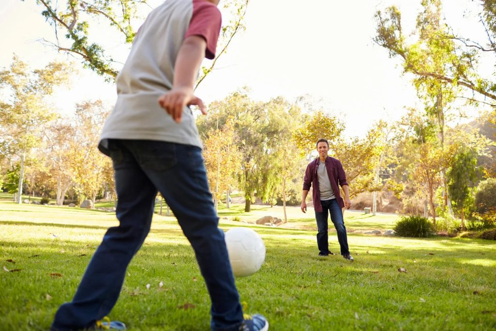 A man in white shirt and jeans kicks a softball to another man in a park