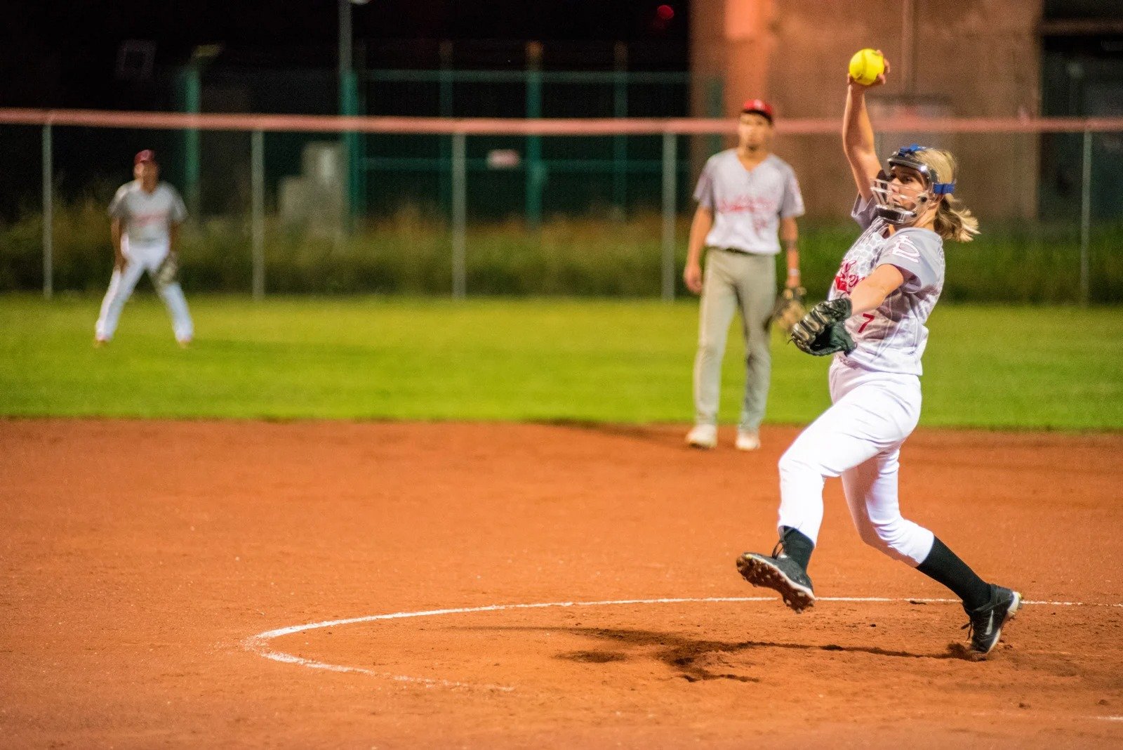 A pitch on a late night softball game field lit up with stadium lights